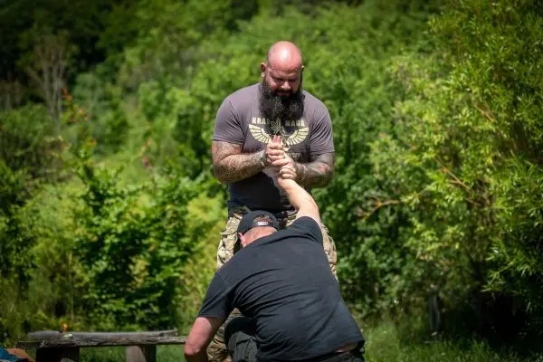 Zwei Personen üben einen Griff im Freien, umgeben von Bäumen und Sträuchern