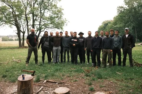 Gruppe von Personen steht auf einer Wiese, umgeben von Bäumen und Holzstämmen