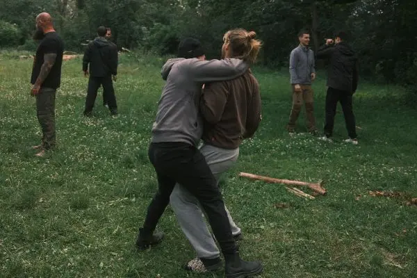 Zwei Personen üben einen Griff im Gras, während andere im Hintergrund stehen