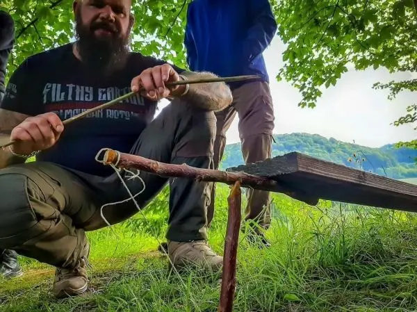 Ein Feuerbohrer wird mit einem Stock auf einem Holzstück bearbeitet