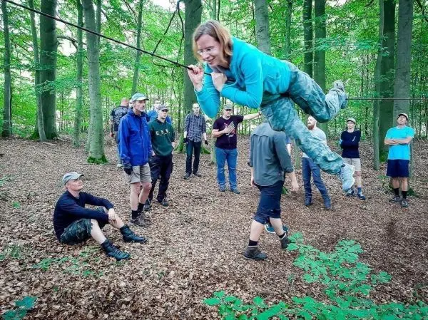 Frau schwingt an Seil über den Boden, umgeben von Zuschauern im Wald