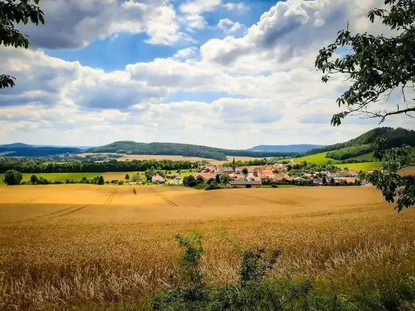 Goldene Felder erstrecken sich bis zu einem kleinen Dorf, umgeben von Hügeln und Wolken