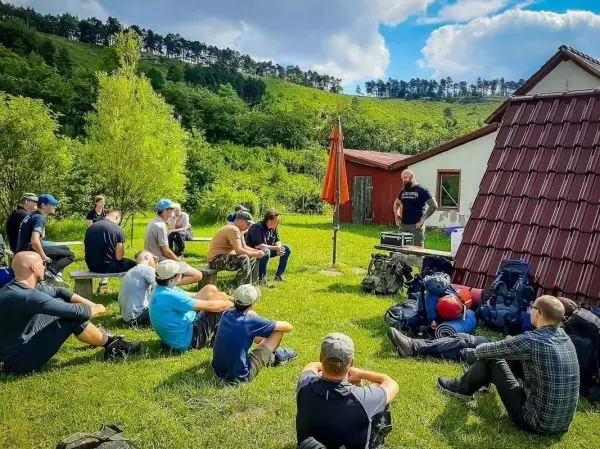 Mann präsentiert Ausrüstung vor sitzender Gruppe auf Wiese neben einem Gebäude