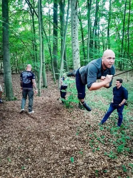Mann springt über einen Hindernis im Wald, während andere zuschauen