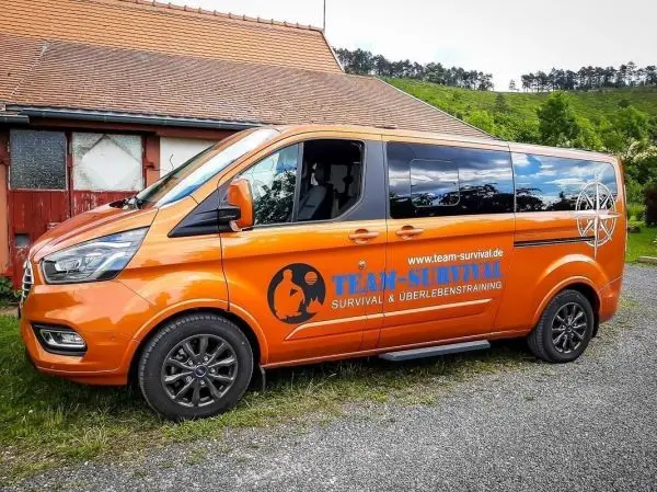 Oranger Transporter mit Logo und Schriftzug für Survival-Training parkt vor einem Gebäude