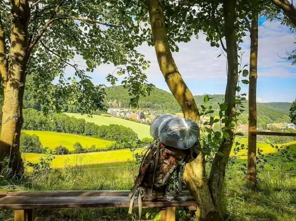 Rucksack lehnt an Baumstamm vor einer grünen Landschaft mit Wiese und Hügeln