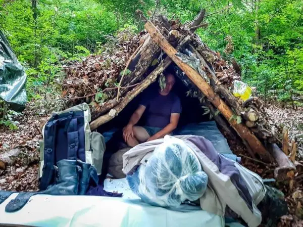 Tarp-Notunterkunft aus Ästen und Laub mit Rucksack und Ausrüstung im Vordergrund