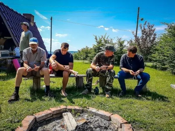 Vier Personen sitzen auf Holzbank vor einem Lagerfeuer, umgeben von Grün