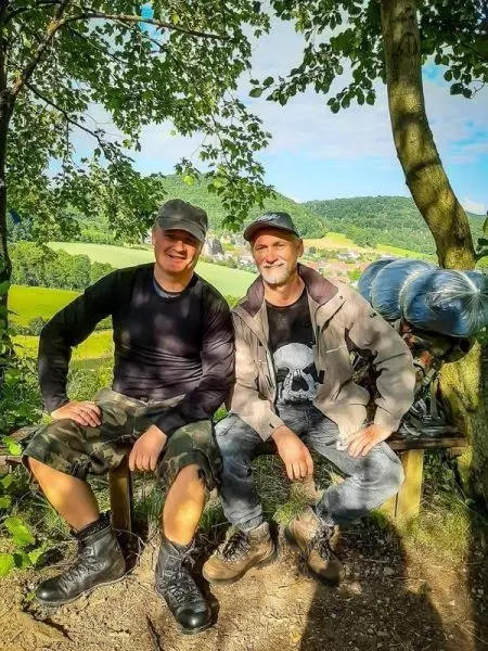 Zwei Personen sitzen auf einer Bank im Freien, im Hintergrund grüne Wiesen und Bäume