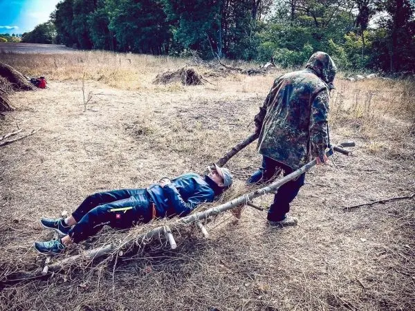 Eine improvisierte Trage aus Ästen transportiert eine Person über den Boden