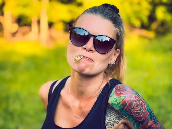 Frau mit Sonnenbrille und tätowiertem Arm hält ein Insekt im Mund