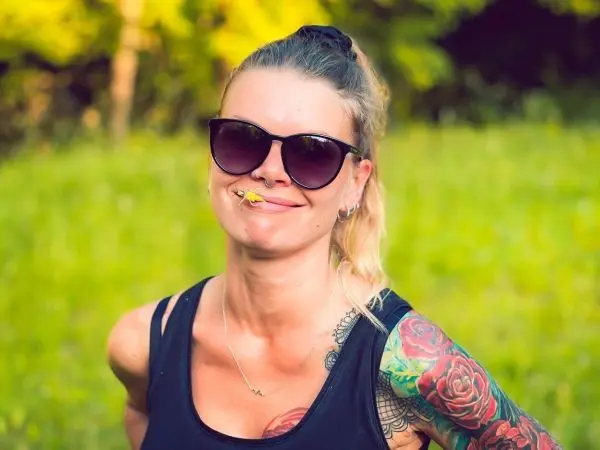 Frau mit Sonnenbrille und Tätowierungen lächelt im Freien mit einem Blatt im Mund