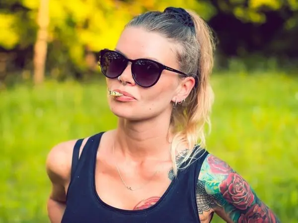 Frau mit Sonnenbrille und Zigarettenstummel im Mund steht in einer grünen Wiese