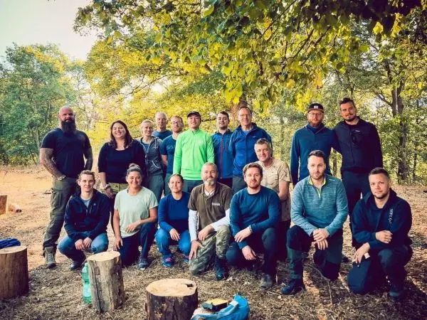 Gruppe von Personen steht vor Bäumen, einige sitzen auf Holzstämmen