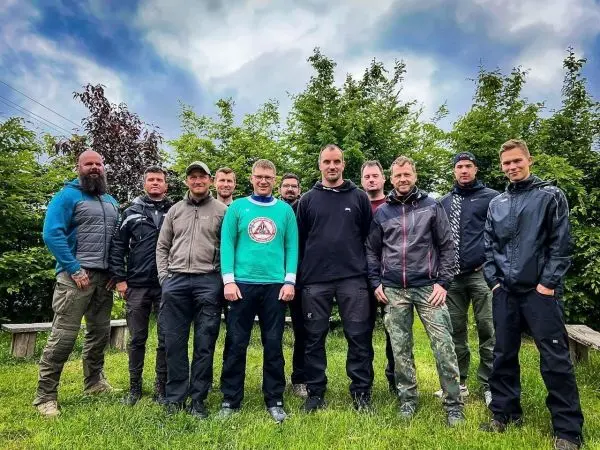 Gruppe von zehn Personen in Outdoor-Bekleidung steht auf einer Wiese vor Bäumen
