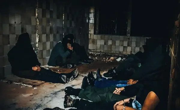 Gruppierung sitzt in einem dunklen Raum auf dem Boden, teilweise mit Kapuzen