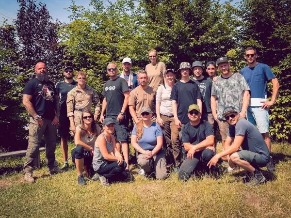 Gruppierung von Personen im Freien vor einer Hecke, einige tragen Outdoor-Bekleidung
