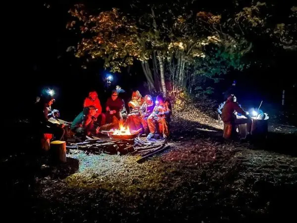 Lagerfeuer umringt von mehreren Personen in der Nacht, mit Stirnlampen beleuchtet