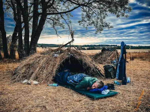 Schlafende Person in einer Laubhütte auf einer Wiese mit Rucksack und Ausrüstung
