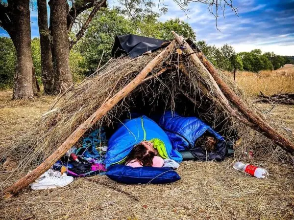 Tarp-Notunterkunft aus Ästen und Gras mit Schlafsäcken im Inneren
