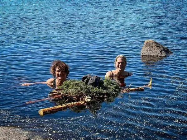 Zwei Personen schwimmen im Wasser neben einer schwimmenden Holz- und Pflanzenkonstruktion
