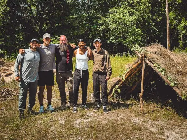 Fünf Personen stehen vor einer selbstgebauten Tarp-Notunterkunft im Freien
