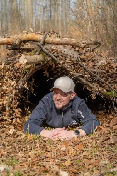 Mann liegt vor einer Laubhütte aus Ästen und Laub im Wald