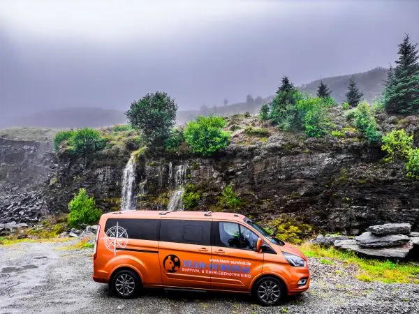 Oranger Transporter steht vor einer Felswand mit Wasserfall und bewaldetem Hang