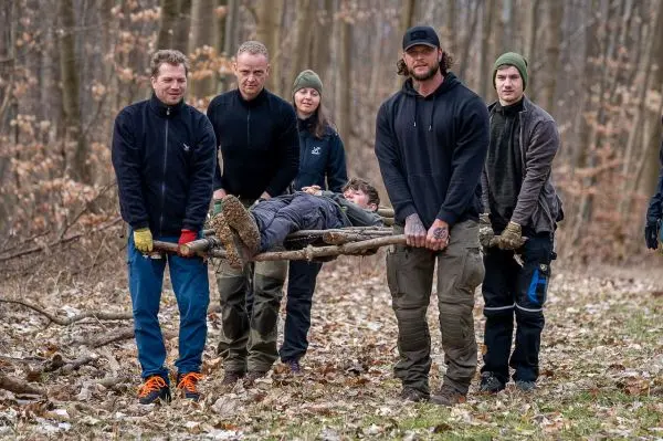 Sechs Personen tragen eine improvisierte Trage aus Ästen mit einer Person darauf