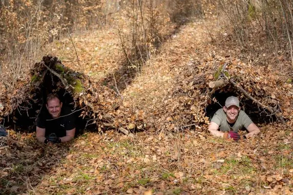 Zwei Personen liegen in improvisierten Laubhütten aus Ästen und Blättern