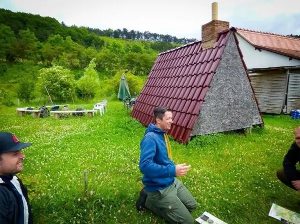 Mann kniet auf Wiese neben einer Holzhütte mit rotem Dach und rauchendem Schornstein