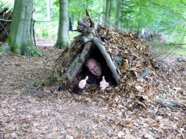 Mann zeigt Daumen hoch aus einer Laubhütte im Wald