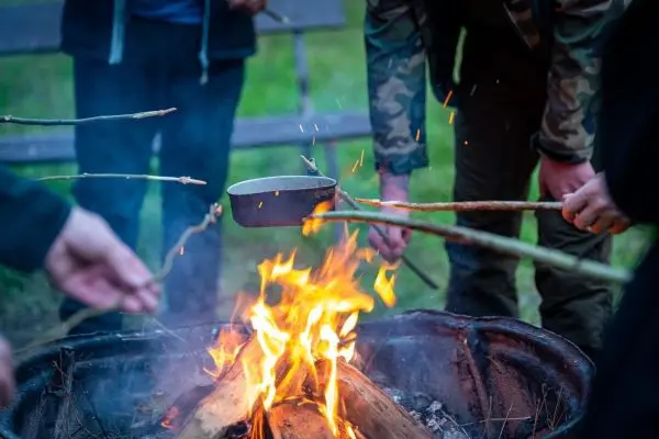 Ein Kessel hängt über einem Lagerfeuer, während mehrere Hände ihn mit Stöcken halten