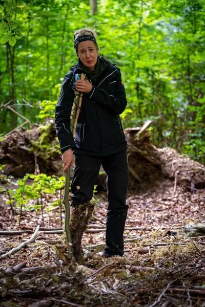 Frau hält einen Stock und steht in einem Waldgebiet mit Laub und Bäumen