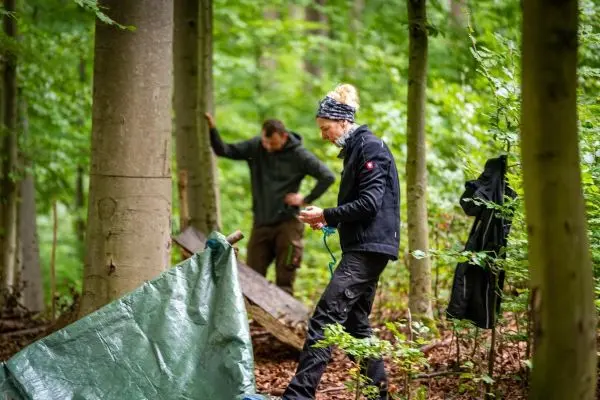 Frau spannt Tarp-Notunterkunft zwischen Bäumen, Mann prüft Holzunterlage