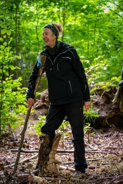 Frau steht mit einem Stock in einem Wald und lächelt