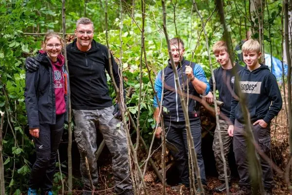 Gruppe von fünf Personen steht im Wald vor einer selbstgebauten Unterkunft