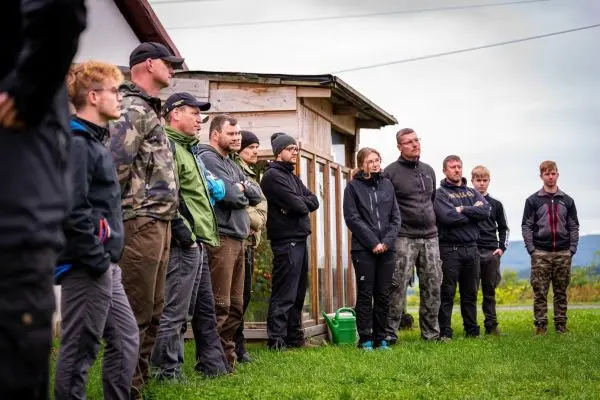 Gruppe von Personen steht in Reihen vor einem Holzgebäude auf einer Wiese