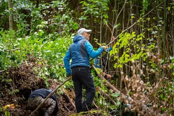 Mann mit blauer Jacke sammelt Äste zur Konstruktion einer Notunterkunft im Wald