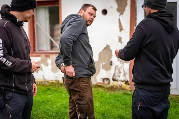 Mann mit Handschellen wird von zwei Personen in der Nähe einer Wand beobachtet