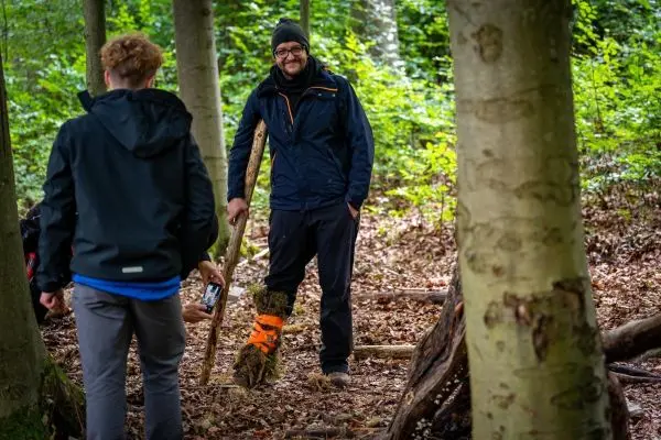 Mann steht mit einem Stock im Wald, während ein anderer ihn filmt