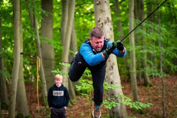 Mann zieht sich an einem Seil zwischen Bäumen entlang, während ein Junge zuschaut