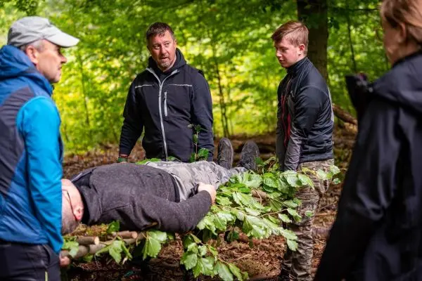 Vier Personen heben eine improvisierte Trage aus Ästen und Blättern