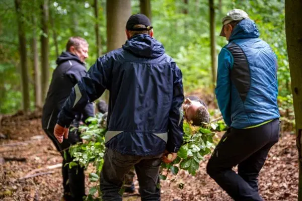 Vier Personen tragen eine improvisierte Trage aus Ästen durch den Wald
