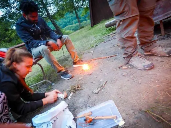 Frau entzündet Zunder mit Feuerstahl, während zwei Personen im Hintergrund sitzen