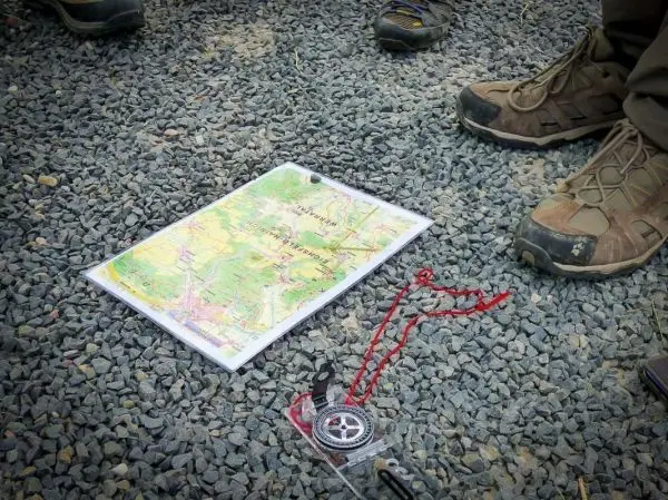 Karte und Kompass liegen auf Kiesboden, Stiefel stehen daneben