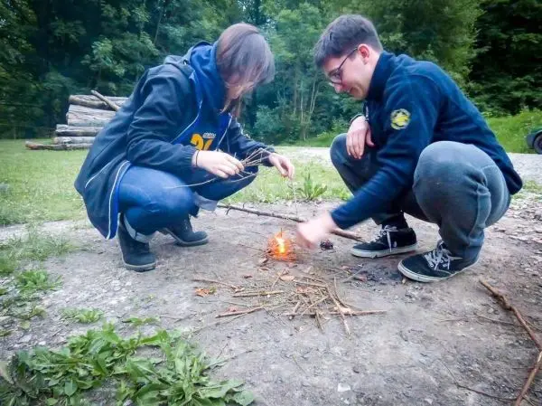Zunder wird mit einem Feuerstahl entzündet, während zwei Personen daneben sitzen