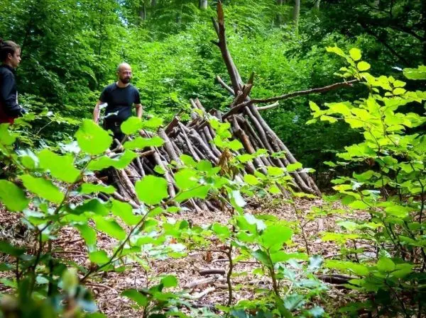 Zwei Personen stehen neben einer selbstgebauten Laubhütte aus Ästen im Wald