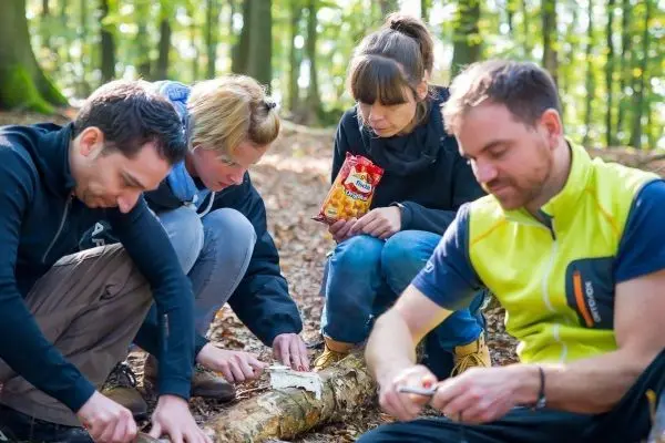 Drei Personen schnitzen mit Messern an einem Baumstamm im Wald