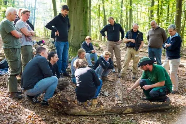 Ein Trainer demonstriert Techniken auf einem Baumstamm, während Teilnehmer aufmerksam zuschauen